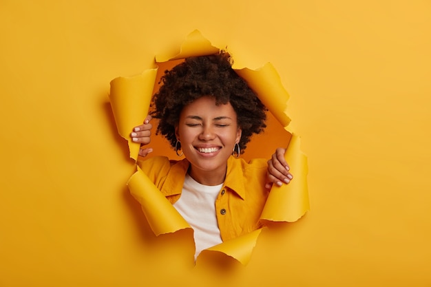 Lucky overjoyed African American woman smiles broadly, has carefree mood, dressed in fashionable clothes poses in yellow paper background