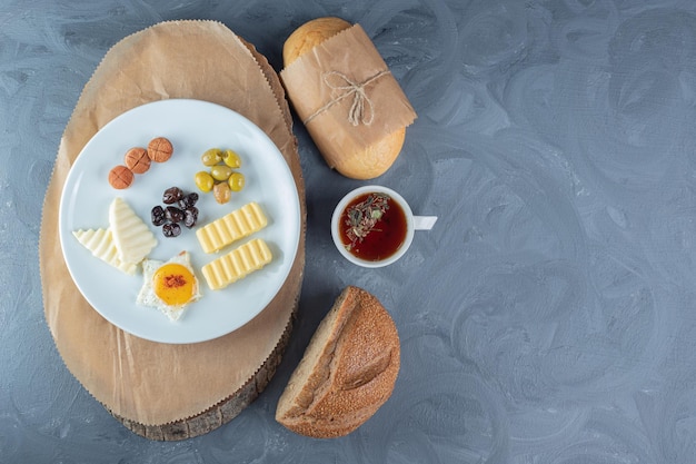 Free photo lumps of bread next to a cup of tea, a glass of juice, and a platter of cheese, egg, butter and sausage slices on a wooden board on marble table.