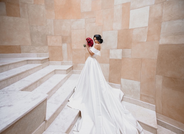 Free photo luxury caucasian brunette bride is standing on the stairs near the stone wall and holding red peonies wedding bouquet