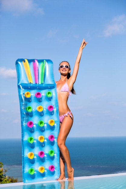 Free photo luxury living, vacation style summer portrait of happy young woman with tanned slim fit body, having sun at luxury villa, holding air mattresses on her hands.