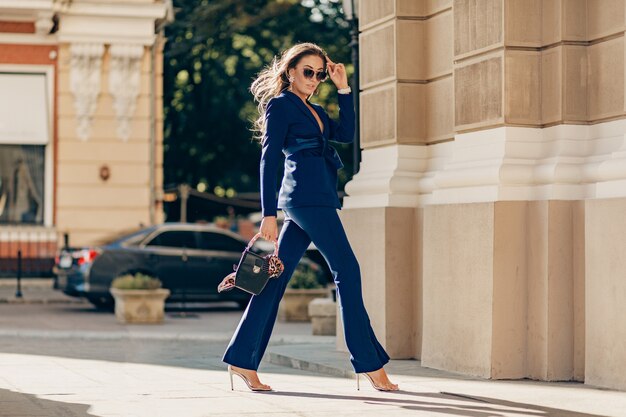Роскошная богатая женщина, одетая в элегантный стильный синий костюм, гуляет по городу в солнечный осенний день, держа кошелек