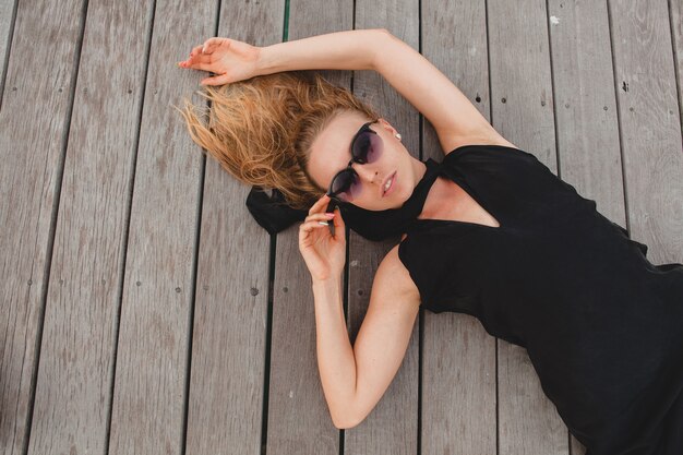 Luxury sexy attractive woman dressed in black dress lying on floor wearing sunglasses, summer vacation, view from above