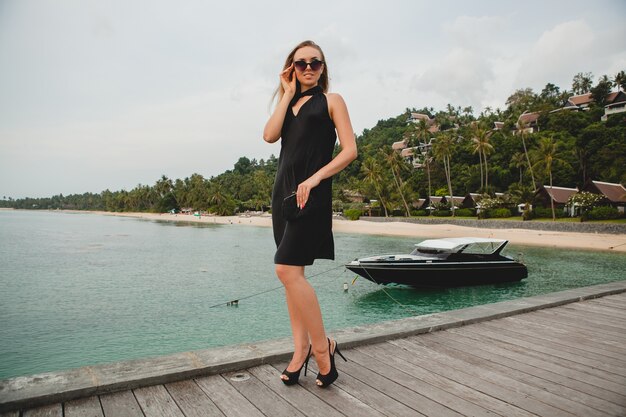 Luxury sexy attractive woman dressed in black dress posing on pier in luxury resort hotel, wearing sunglasses, summer vacation, tropical beach