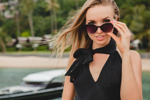 Free photo luxury sexy attractive woman dressed in black dress posing on pier in luxury resort hotel, wearing sunglasses, summer vacation, tropical beach