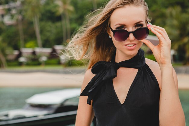 Luxury sexy attractive woman dressed in black dress posing on pier in luxury resort hotel, wearing sunglasses, summer vacation, tropical beach