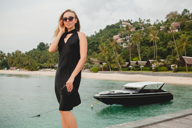 Бесплатное фото Роскошная сексуальная привлекательная женщина, одетая в черное платье, позирует на пирсе в роскошном курортном отеле, в солнцезащитных очках, летние каникулы, тропический пляж
