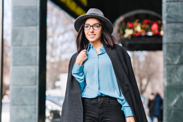 Роскошный стильный городской портрет модной молодой женщины, идущей по улице в городе во время Рождества. Серая шляпа, пальто, волосы брюнетки, черные очки, синяя рубашка, бодрое настроение, бизнесвумен.