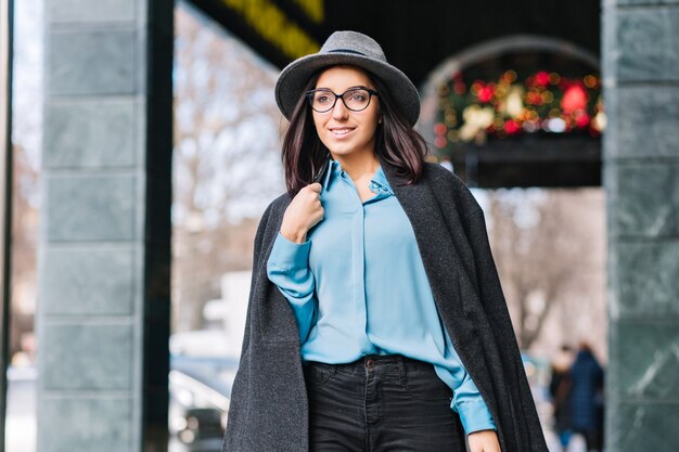 Luxury stylish city portrait fashionable young woman walking on street in city at christmas time. Grey hat, coat, brunette hair, black glasses, blue shirt, cheerful mood, businesswoman.