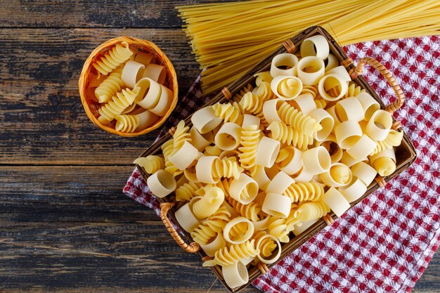 Macaroni pasta in a basket and bucket with spaghetti top view on a picnic cloth and wooden background