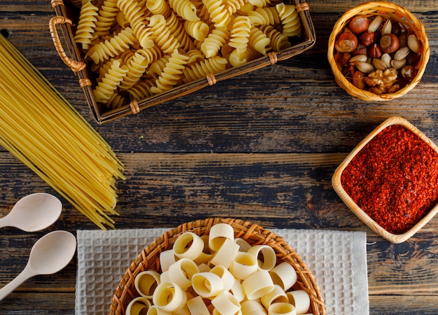 Macaroni pasta in a basket with spaghetti, spoons, various nuts top view on a wooden background space for text