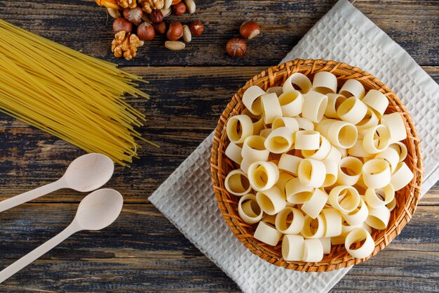 Macaroni pasta in a basket with spaghetti, spoons, various nuts top view on a wooden background