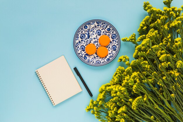 Macaroons on ceramic plate; spiral notepad; pen and bunch of yellow flowers on blue background