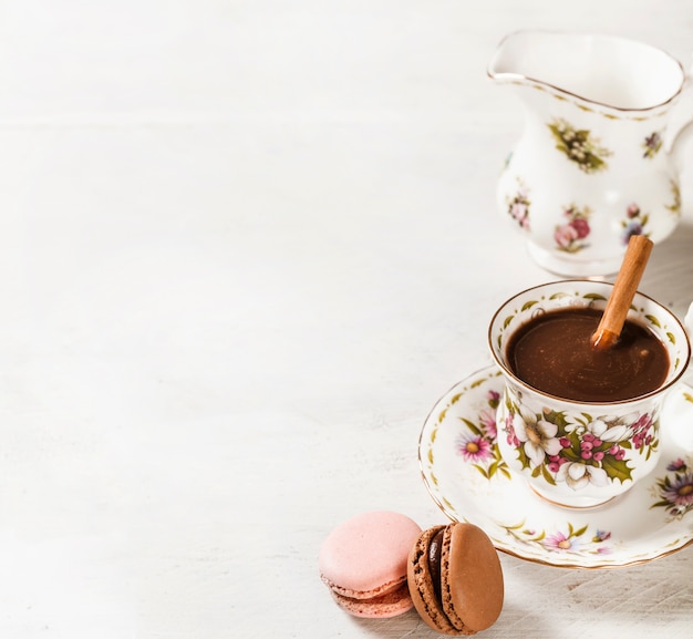 Macaroons and hot chocolate with cinnamon stick in ceramic cup on white textured backdrop