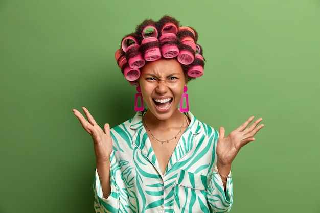 Free photo mad angry housewife gestures with annoyance, screams at husband or children, expresses irritation, feels sick and tired of domestic work, demands time for herself, makes hairstyle, wears curlers