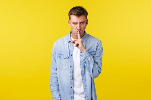 Mad and annoyed serious-looking blond man tell be quiet, shushing at camera with bothered angry face, asking keep quiet, be silent as standing over yellow background.