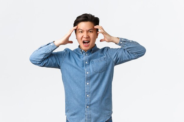 Mad and distressed asian man looking furious and frustrated, losing temper, holding hands on head and frowning angry, losing control over emotions, feeling anger and rage, white background.