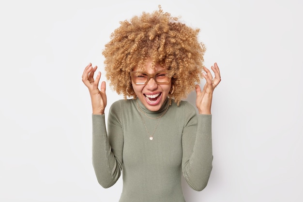 Mad emotional overemotive woman with curly hair screams loudly shakes hands near head keeps mouth opened feels very happy wears spectacles and casual poloneck isolated over white background.