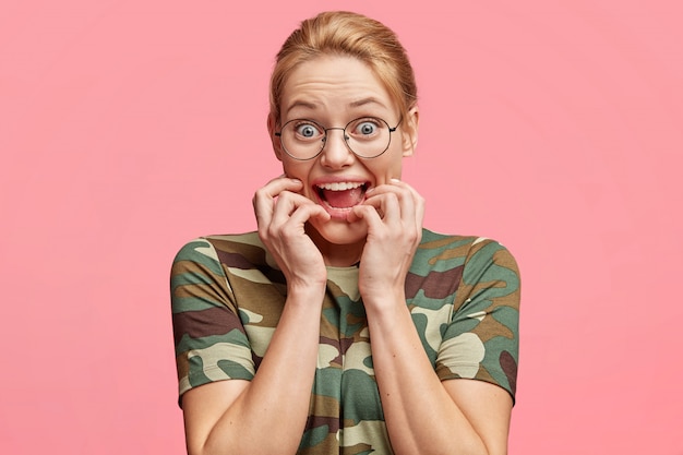 Mad surprised female model with eyes popped out, stares through round spectacles, bites finger nails, feels nervous and excited, reacts on something very actively, isolated over pink