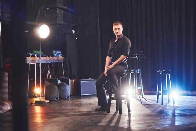 Magic look. Handsome young man in black clothes sitting on chair near in dark room with light.