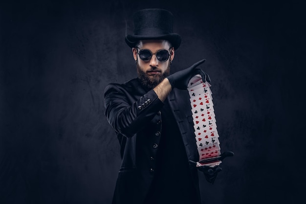 Magician in a black suit, sunglasses and top hat, showing trick with playing cards on a dark background.