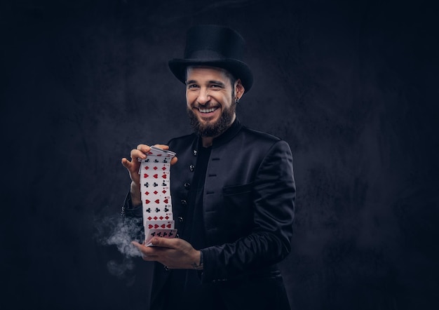 Free photo magician in a black suit and top hat, showing trick with playing cards and magic smoke on a dark background.