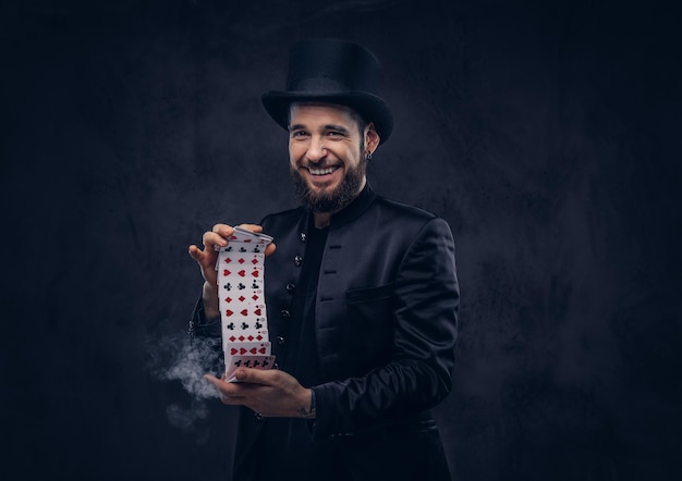 Фокусник в черном костюме и цилиндре, демонстрирующий трюк с игральными картами и волшебным дымом на темном фоне.