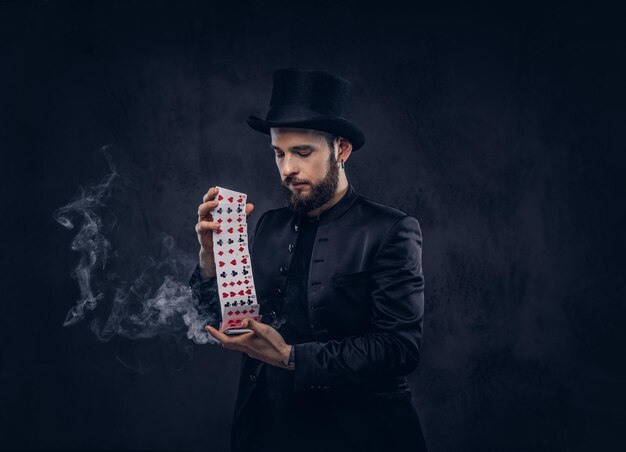 Magician in a black suit and top hat, showing trick with playing cards and magic smoke on a dark background.