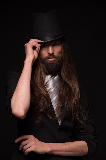 Free photo magician with moustache touching histop hat. long-haired man in black suit isolated on dark background.