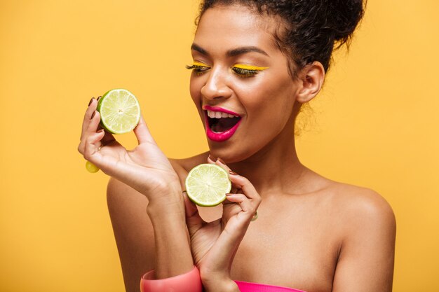 magnificent african american woman with trendy makeup holding two halves of fresh lime in both hands isolated, over yellow wall