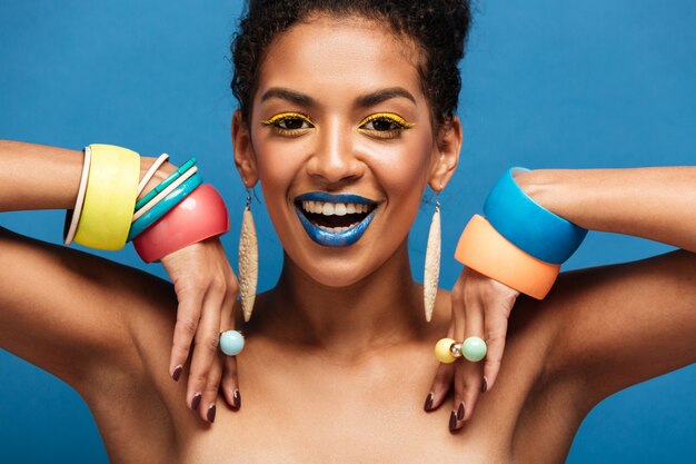 magnificent half-naked woman with colorful makeup smiling and demonstrating accessories on her arms, isolated over blue