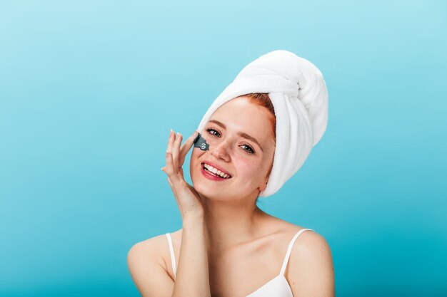 Magnificent young woman applying face mask. Studio shot of inspired girl doing skincare treatment with smile on blue background.