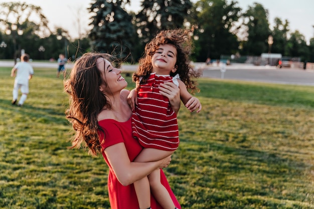 Magnificent young woman carrying her daughter and smiling. Charming lady looking with love at curly kid.