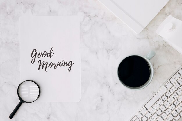 Magnifying glass on good morning paper with coffee cup; diary and keyboard on white marble desk