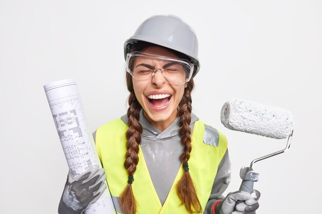 유지 보수 및 직업 개념. 기뻐하는 여성 엔지니어가 흰 벽 위에 절연 건설 현장에서 재건 바쁜 안전 옷을 입은 청사진 및 페인트 롤러로 포즈