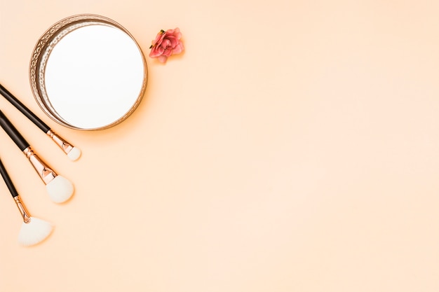 Кисточки для макияжа; круглое зеркало и роза на бежевом фоне