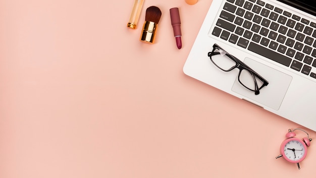 Free photo makeup brush,lipstick near the laptop with eyeglasses and alarm clock on colored background