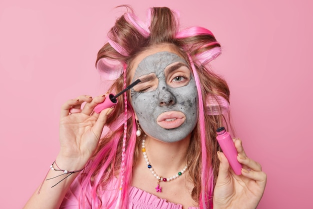Бесплатное фото Макияж визаж и концепция ухода за кожей прекрасная молодая европейская женщина наносит косметическую глиняную маску, наносит тушь, бигуди для волос, готовится к свиданию, подмигивает, носит ожерелье, изолированное на розовом фоне