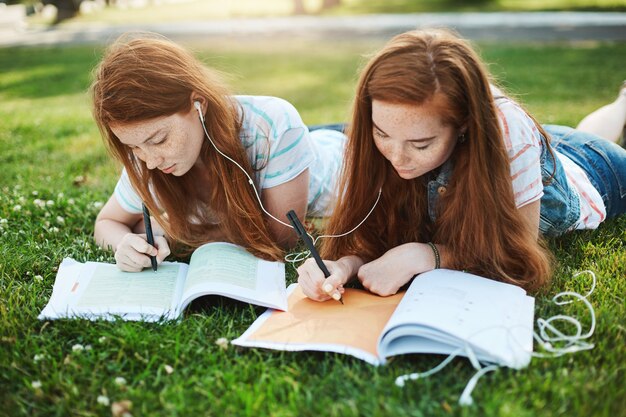 Making homework can be fun. Outdoor shot of two attractive redhead girls with freckles, lying on grass in park, sharing earphones and writing essays for university on fresh air, helping each other.
