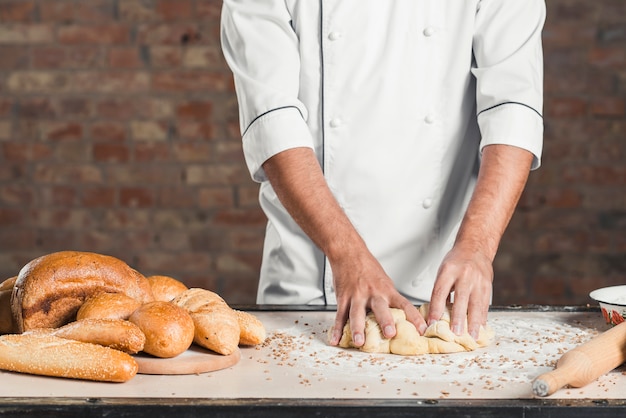 Мужской пекарь замешивает тесто на кухонной столешнице со многими хлебами