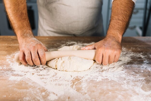 Male baker's hand flattening dough with rolling pin on wooden table