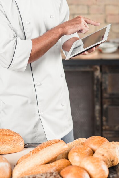 Male baker standing behind the table with breads using digital tablet