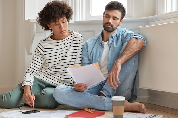 Male bearded accountant and his secretary work together in modern apartment, pose on wooden floor and discuss financial report, have serious gaze in papers, study analytics, feel comfort at home