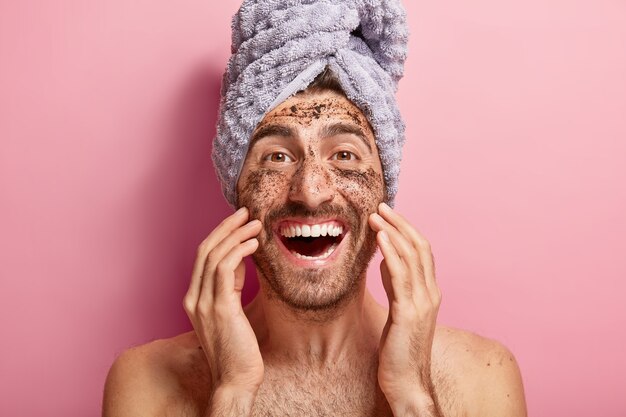 Male beauty concept. Happy joyous man applies coffee scrub on face, removes dark dotes, wants to look refreshed, has wrapped towel on head