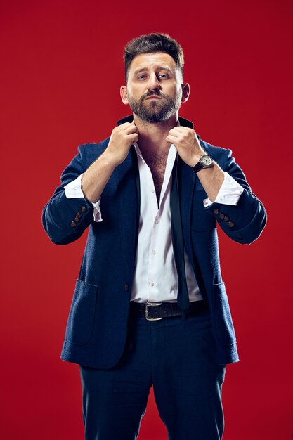 Male beauty concept. Portrait of a fashionable young man with stylish haircut wearing trendy suit posing over red background. Perfect hair. Elegant italian style.