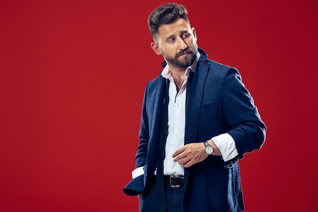 Male beauty concept. Portrait of a fashionable young man with stylish haircut wearing trendy suit posing over red background.