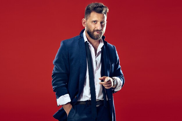 Male beauty concept portrait of a fashionable young man with stylish haircut wearing trendy suit posing over red wall