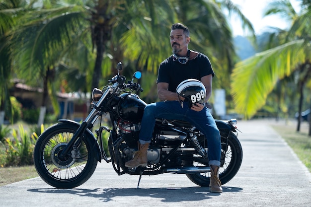 Male biker with a thick beard poses on a motorbike in a beautiful park