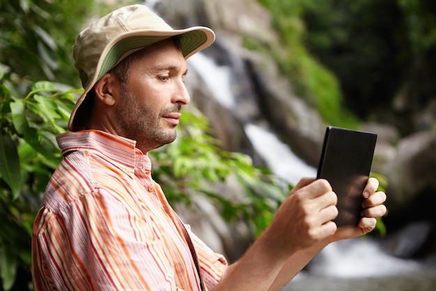 Male biologist in striped shirt and hat working in nature park, taking picture or recording video of wildlife using his black digital tablet standing against waterfall and green trees
