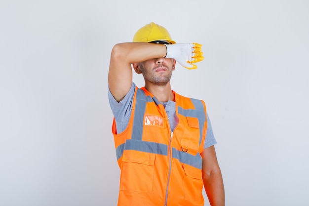 Free photo male builder covering eyes with arm in uniform, helmet, gloves and looking serious and sad, front view.