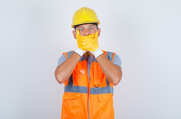 Строитель-мужчина прикрывает рот руками за ошибку в униформе, шлеме, перчатках и выглядит шокированным, вид спереди